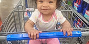 Mckenzie is registered to the contest to win money with this photo: aisle, baby, child, curly_hair, cute, floor, grocery, happy, holding, indoor, metal_cart, person, pink_clothing, retail, shopping, shopping_cart, smiling, socks, store, white_shirt