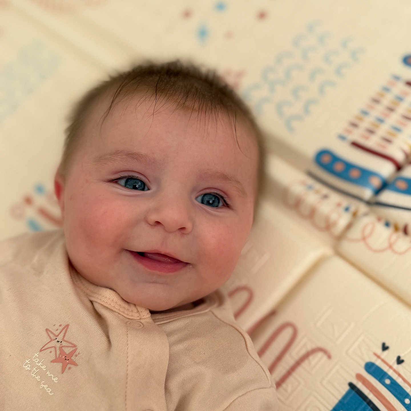 Elena participe au concours pour gagner de l'argent avec cette photo : adorable, baby, blue_eyes, cheeks, child, closeup, clothing, cute, face, happy, indoor, infant, lying_down, onesie, patterned_background, play_mat, portrait, skin, smiling, soft_focus