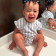 Amina is registered to the contest to win money with this photo: baby, child, smiling, laughing, mirror, reflection, blue_hair_ties, curly_hair, striped_clothing, white_surface, indoor, cute, happy, sitting, portrait, skin, hand, foot, face, expression