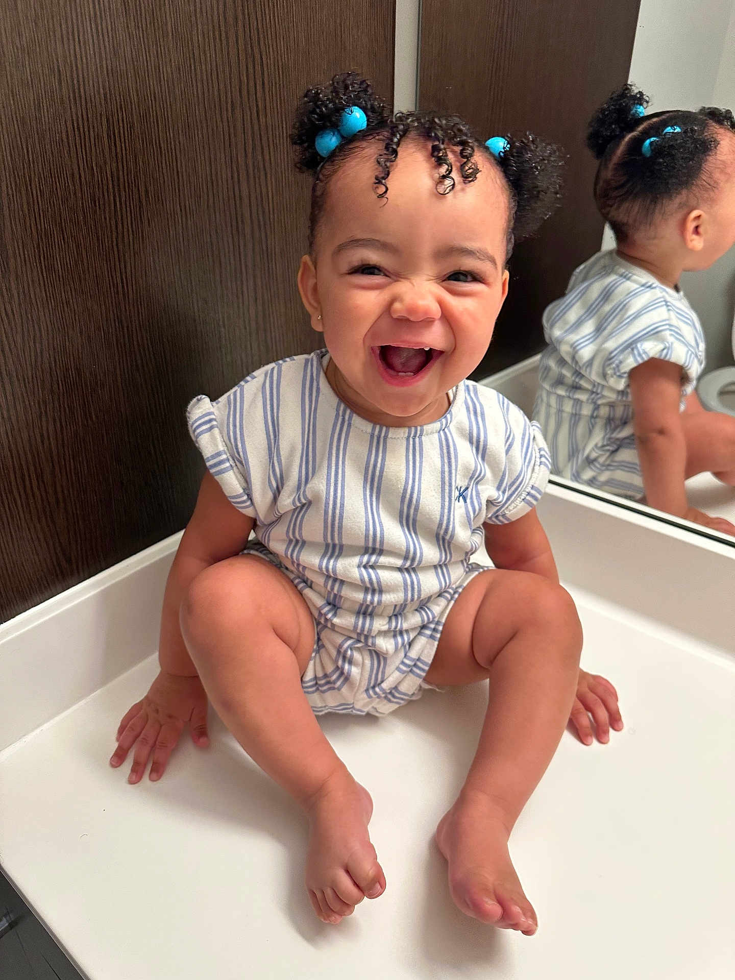 Amina is registered to the contest to win money with this photo: baby, child, smiling, laughing, mirror, reflection, blue_hair_ties, curly_hair, striped_clothing, white_surface, indoor, cute, happy, sitting, portrait, skin, hand, foot, face, expression