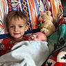 child, baby, blanket, teddy_bear, smile, sleeping, couch, plush_toy, patterned_blanket, red_clothing, cozy, indoor, family, love, portrait, young_child, infant, comfort, toddler, resting