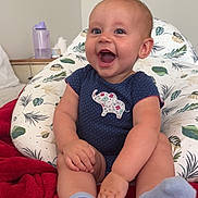 Octavia is registered to the contest to win money with this photo: baby, smile, onesie, blue_socks, red_blanket, cushion, leaf_pattern, indoor, happy, child, person, sitting, cute, infant, face, hand, footwear, room, background, expression