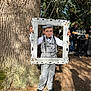 child, boy, vintage_clothing, checkered_suit, bow_tie, suspenders, newsboy_cap, picture_frame, tree, outdoor, sunlight, shadow, people, food_station, event, grass, nature, portrait, smile, casual