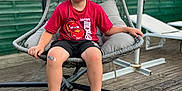 Léonardo participe au concours pour gagner de l'argent avec cette photo : child, boy, sitting, hanging_chair, outdoor, red_shirt, lightning_mcqueen, bandage, knee, sneakers, wooden_deck, fence, pillow, smile, casual_clothing, daytime, relaxing, playful, summer, young