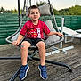 child, boy, sitting, hanging_chair, outdoor, red_shirt, lightning_mcqueen, bandage, knee, sneakers, wooden_deck, fence, pillow, smile, casual_clothing, daytime, relaxing, playful, summer, young
