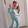 child, girl, pigtails, unicorn, sweatshirt, pants, white_shoes, pose, hand_sign, indoor, door, floor, smile, hair, casual_clothing, cute, playful, young, person, confident