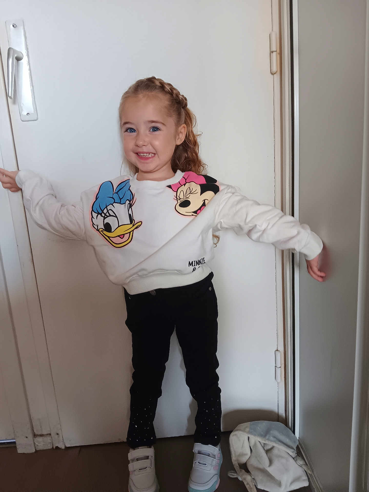 Chloé a rejoint le concours — aidez-le/la à gagner de superbes lots ! child, girl, smiling, braided_hair, sweatshirt, donald_duck, minnie_mouse, black_pants, white_sneakers, indoor, door, bag, standing, happy, pose, clothing, footwear, casual, person, portrait