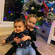 Chloé a rejoint le concours — aidez-le/la à gagner de superbes lots ! child, children, baby, smiling, hugging, christmas_tree, holiday, indoor, floor, decorations, ornaments, lights, festive, joyful, family, toddler, jeans, long_sleeve_shirt, window, home