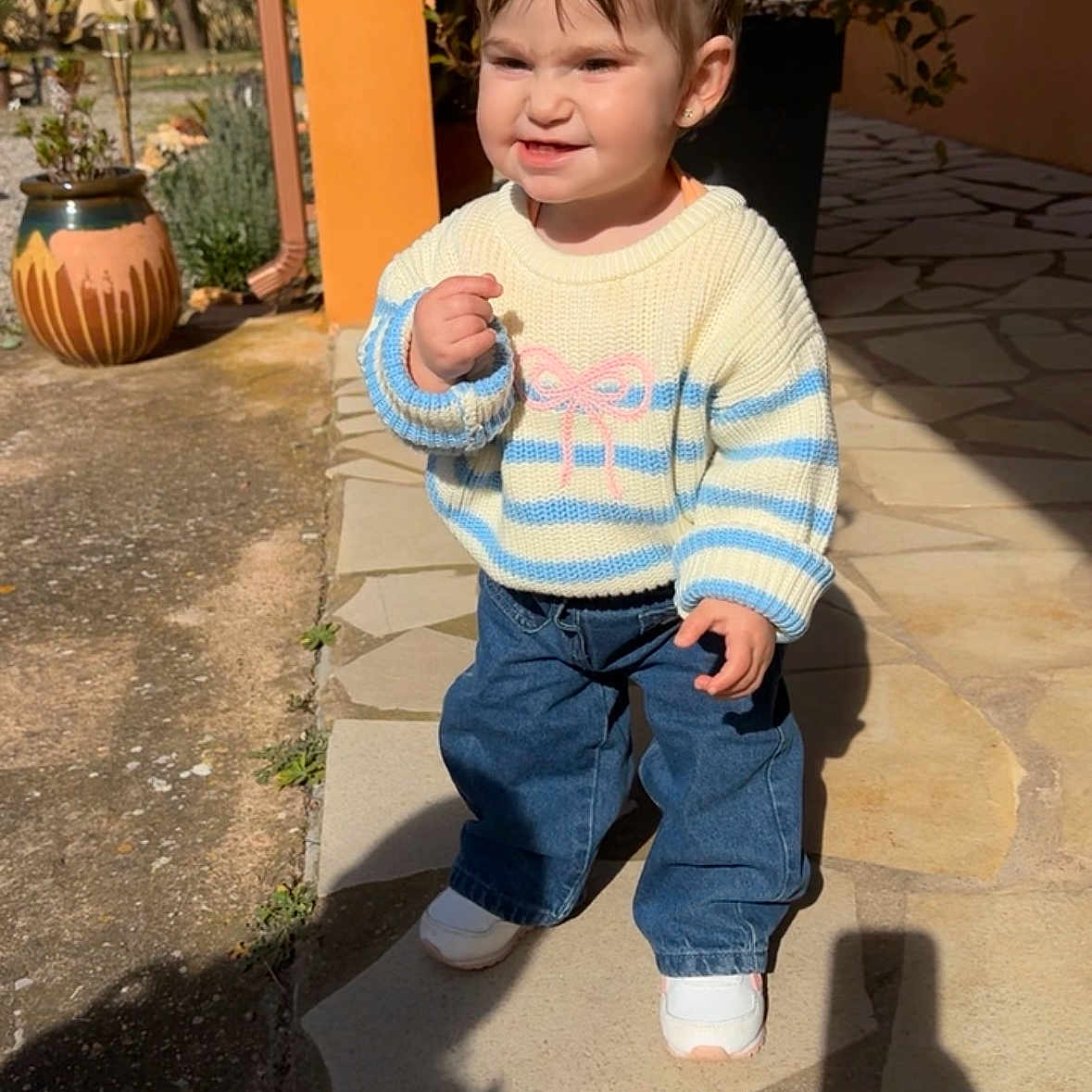 Vicenta a rejoint le concours — aidez-le/la à gagner de superbes lots ! building, child, cute, daylight, determined_face, expression, jeans, outdoor, person, pigtails, plant, pottery, shadow, sidewalk, striped_clothing, sunlight, sweater, toddler, white_shoes, window