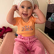 Vicenta a rejoint le concours — aidez-le/la à gagner de superbes lots ! basket, bed, child, clothing, cute, face, furniture, happy, headwear, indoor, orange_shirt, person, pink_pants, playful, room, sitting, smiling, toddler, towel, young_child