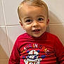 toddler, child, smiling, red_shirt, graphic_tshirt, astronaut_bear, indoor, portrait, face, happy, blonde_hair, short_hair, cute, sitting, casual_clothing, young_child, person, tile_wall, playful, bright_colors