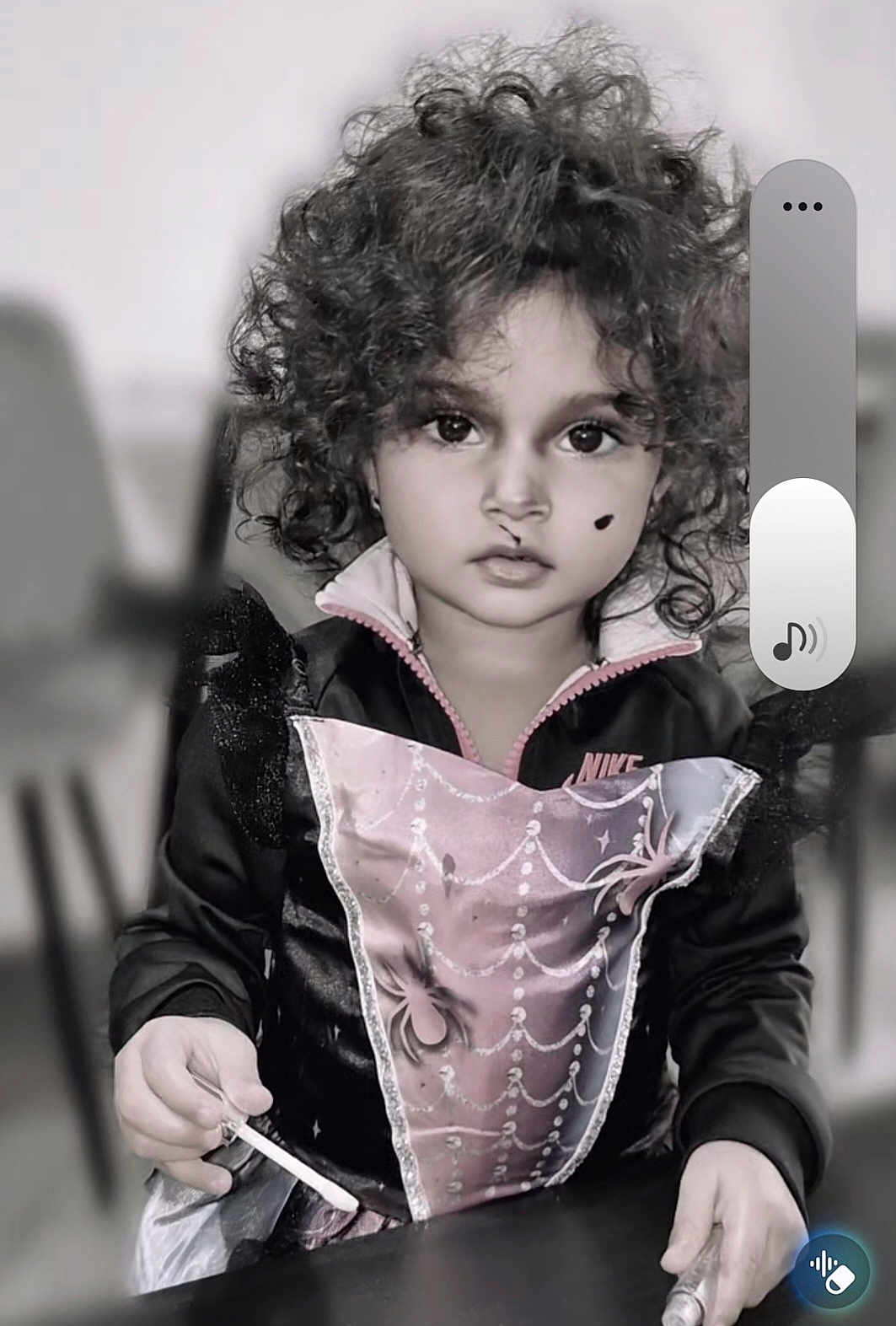 Mayle is registered to the contest to win money with this photo: child, curly_hair, dress, nike_jacket, face, hands, table, chair, indoor, portrait, black_and_white, beauty_mark, spider_web_pattern, costume, young, person, curious, holding, sitting, casual