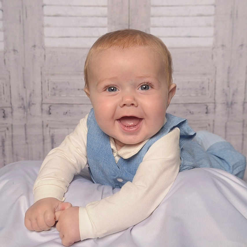 Peter-Philip is registered to the contest to win money with this photo: baby, background, blanket, blue_vest, child, clothing, cream_shirt, cute, face, happy, indoors, infant, lying_down, person, portrait, smiling, soft_lighting, studio_photo, toddler, wooden_wall