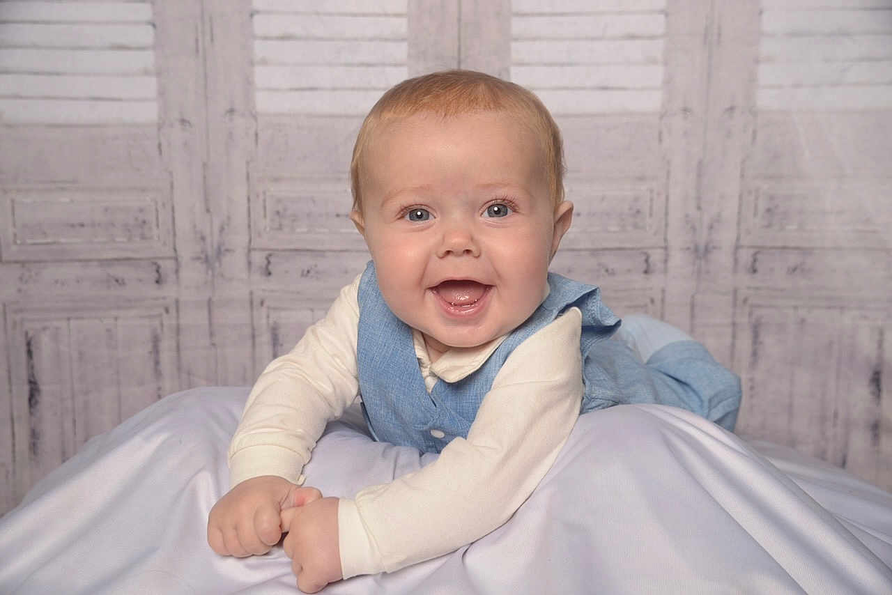 Peter-Philip is registered to the contest to win money with this photo: baby, background, blanket, blue_vest, child, clothing, cream_shirt, cute, face, happy, indoors, infant, lying_down, person, portrait, smiling, soft_lighting, studio_photo, toddler, wooden_wall