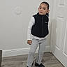 child, portrait, standing, indoors, hallway, door, wood_floor, fashion, vest, sweatpants, long_sleeves, shoes, serious_expression, curly_hair, full_body, home, wall, baseboard, casual_clothing, photography