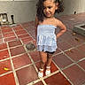 child, girl, toddler, curly_hair, dress, white_outfit, sandals, standing, portrait, outdoor, patio, tiles, pavement, fence, wall, leaves, sunlight, fashion, confident_pose, smiling
