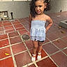 child, girl, toddler, portrait, outdoor, patio, standing, smiling, curly_hair, hairstyle, dress, sparkly_clothing, sandals, fashion, confident, posing, courtyard, tiles, fence, daylight