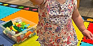 Jade is registered to the contest to win money with this photo: toddler, child, toy, play_mat, indoor, colorful, building_blocks, shoes, shorts, floral_shirt, person, floor, playroom, furniture, plastic_container, carpet, footwear, hand, expression, young_child