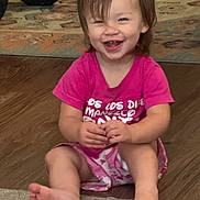 Jade is registered to the contest to win money with this photo: toddler, child, smiling, sitting, indoors, pink_shirt, shorts, barefoot, happy, face, hair, floor, wood_floor, rug, toy_wheel, person, cute, young_child, expression, casual_clothing