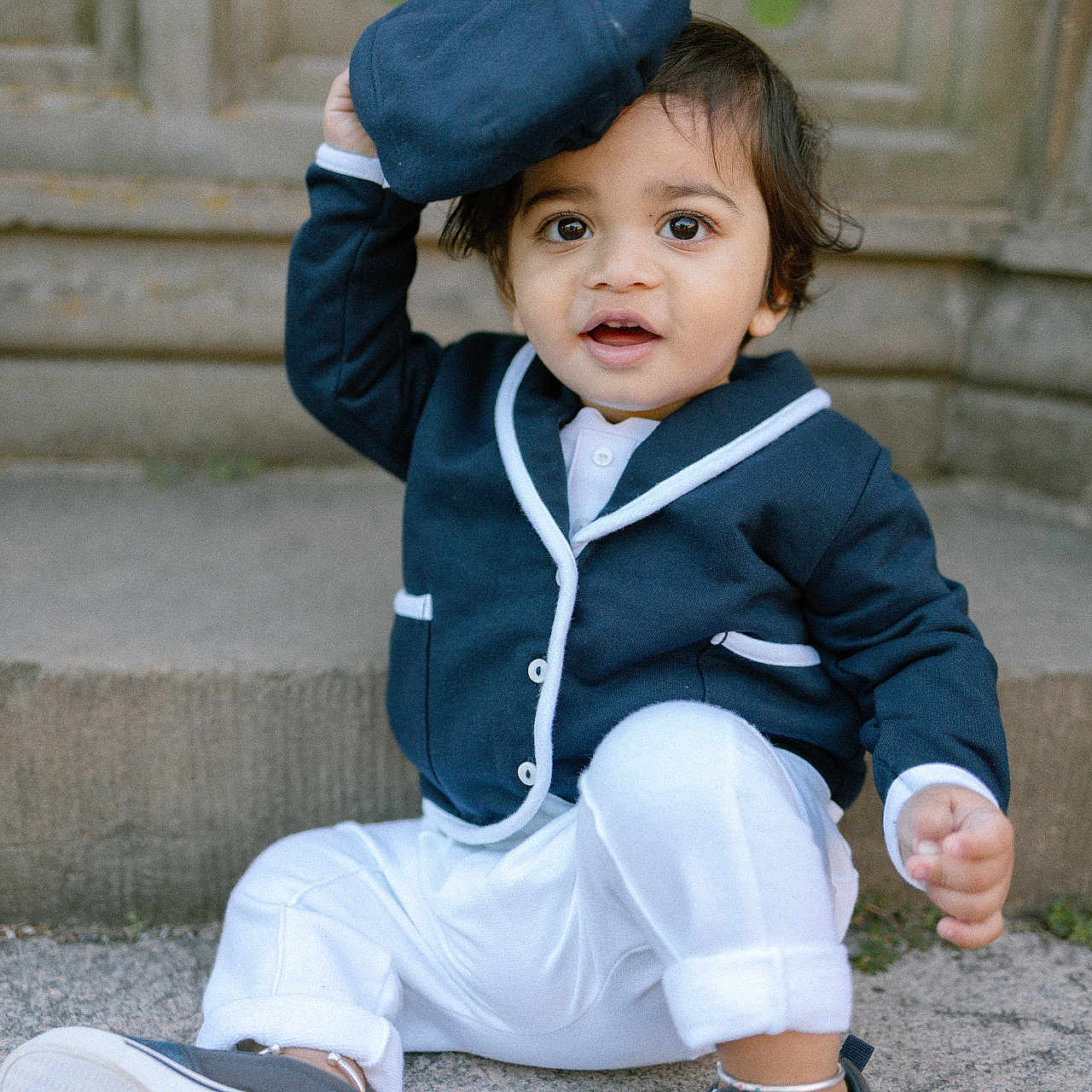 Krishiv joined the competition — help win amazing prizes! toddler, child, baby, blue_clothing, white_pants, navy_cap, sneakers, sitting, outdoor, stone_background, curly_hair, cute, playful, portrait, happy, person, casual, young_child, smiling, fashion
