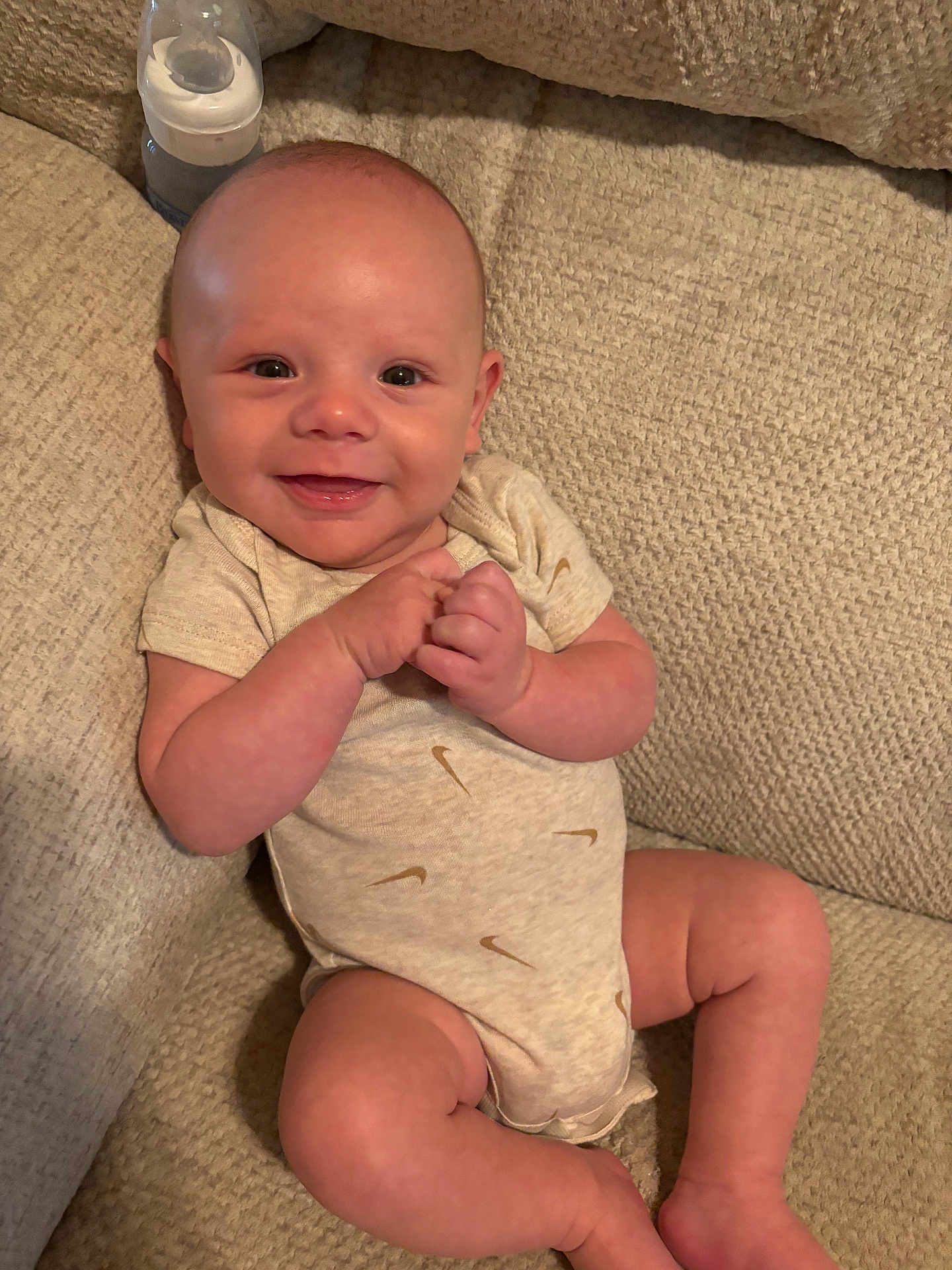 Grayson is registered to the contest to win money with this photo: baby, smiling, onesie, sitting, couch, bottle, happy, infant, face, hands, legs, beige, texture, clothing, person, indoor, cute, portrait, child, relaxed