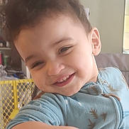 Donovan is registered to the contest to win money with this photo: toddler, child, smiling, curly_hair, blue_outfit, indoor, baby_gate, furniture, home, playful, person, casual, young_child, happy, face, arm, table, cozy, portrait, candid