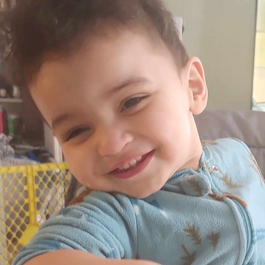 Donovan is registered to the contest to win money with this photo: toddler, child, smiling, curly_hair, blue_outfit, indoor, baby_gate, furniture, home, playful, person, casual, young_child, happy, face, arm, table, cozy, portrait, candid