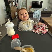 Pierre participe au concours pour gagner de l'argent avec cette photo : baby, baby_food, bottle, cabinet, child, cloth, couch, curtain, feeding, floor, high_chair, indoor, living_room, person, plant, smiling, spoon, table, television, window