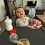 baby, baby_food, bottle, cabinet, child, cloth, couch, curtain, feeding, floor, high_chair, indoor, living_room, person, plant, smiling, spoon, table, television, window