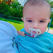 David is registered to the contest to win money with this photo: baby, pacifier, child, blue_clothing, outdoor, park, grass, trees, person, closeup, face, head, infant, daylight, nature, holding, serious, portrait, cute, young