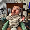 baby, child, clock, clothing, curious, eyes, face, furniture, hand, high_chair, indoor, infant, living_room, portrait, sitting, smiling, striped_shirt, television, toys, window_blinds