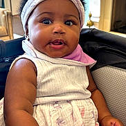 Logan is registered to the contest to win money with this photo: baby, child, headband, dress, indoor, curly_hair, arm, hand, teething_toy, seated, face, skin, furniture, lamp, fireplace, comfort, expression, portrait, closeup, cute