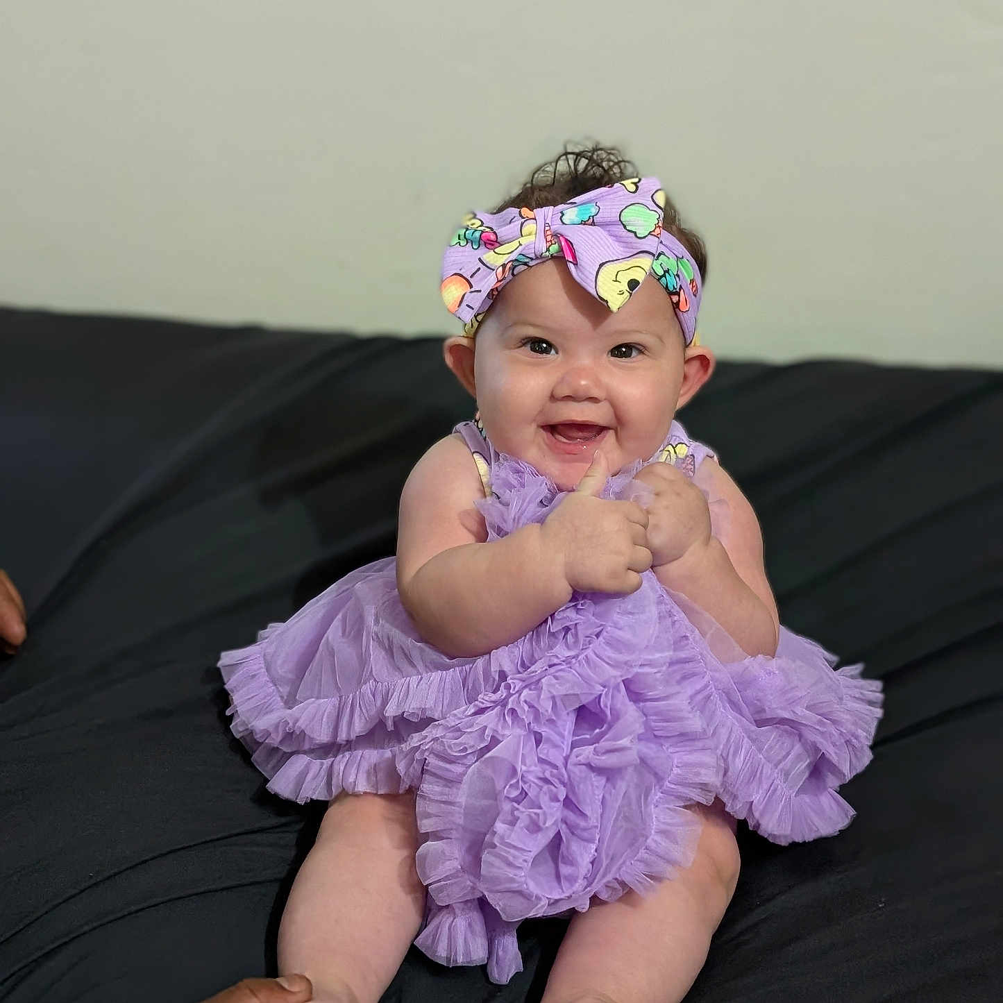 Angélique is registered to the contest to win money with this photo: adult_hand, baby, black_background, child, curly_hair, cute, dress, feet, hands, happy, headband, indoor, infant, person, portrait, purple, sitting, smiling, support, thumbs_up