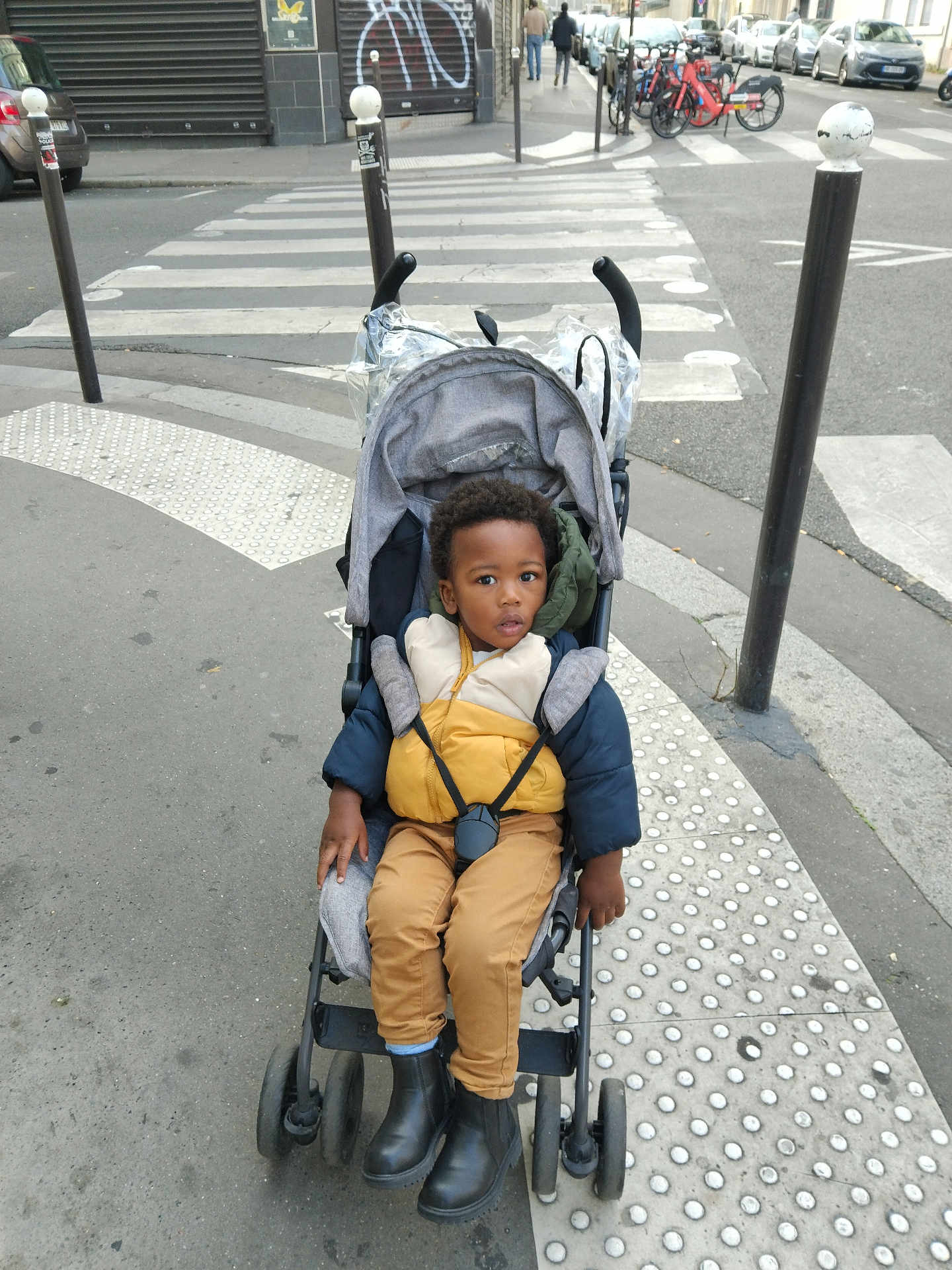 Rodady participe au concours pour gagner de l'argent avec cette photo : toddler, child, stroller, puffer_jacket, boots, sidewalk, urban, crosswalk, pedestrian, bicycle, street, city, outerwear, winter_clothing, curb, metal_pole, asphalt, baby, portrait, outdoor