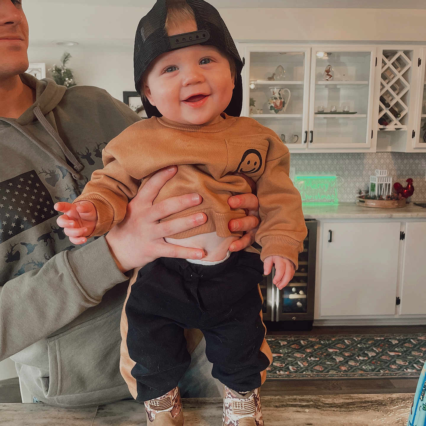 Sawyer is registered to the contest to win money with this photo: adult_hand, appliance, baby, cabinet, cap, casual, child, clothing, counter, decor, face, footwear, happy, holding, indoor, kitchen, person, portrait, smiling, table