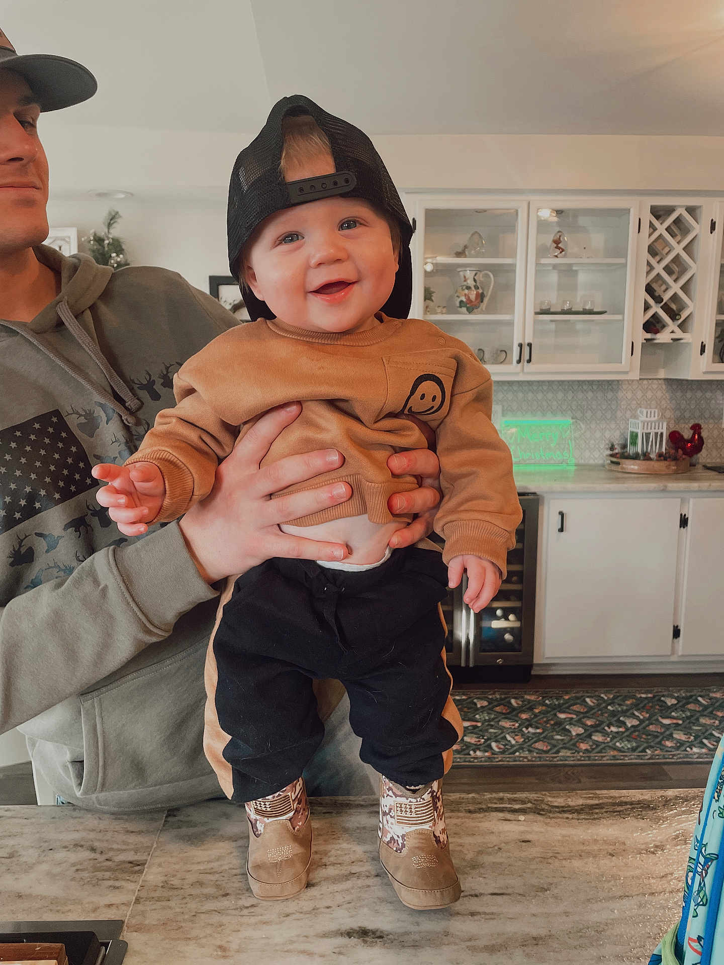 Sawyer is registered to the contest to win money with this photo: baby, child, smiling, cap, adult_hand, kitchen, counter, cabinet, clothing, footwear, indoor, person, happy, face, holding, table, decor, appliance, portrait, casual