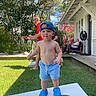 child, toddler, boy, outdoor, backyard, portrait, smiling, baseball_cap, blue_clogs, shirtless, shorts, cornhole, beanbags, lawn, house, porch, trees, sunlight, family, summer