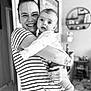 baby, person, smile, striped_shirt, holding, indoor, black_and_white, tattoo, jeans, happy, face, arm, home, portrait, child, furniture, shelf, doorway, floor, cozy