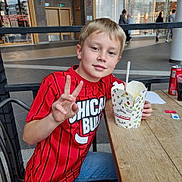 Arthur a rejoint le concours — aidez-le/la à gagner de superbes lots ! child, boy, red_shirt, chicago_bulls, peace_sign, food, takeout_box, spoon, table, wooden_table, indoor, shopping_mall, casual_clothing, smiling, seated, portrait, young_person, light_hair, jeans, public_space