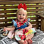 Lila is registered to the contest to win money with this photo: baby, child, smiling, red_bow, dress, toy, wooden_bench, blanket, patterned, blue_eyes, happy, sitting, outdoor, cute, infant, person, hand, feet, colorful_clothing, playful