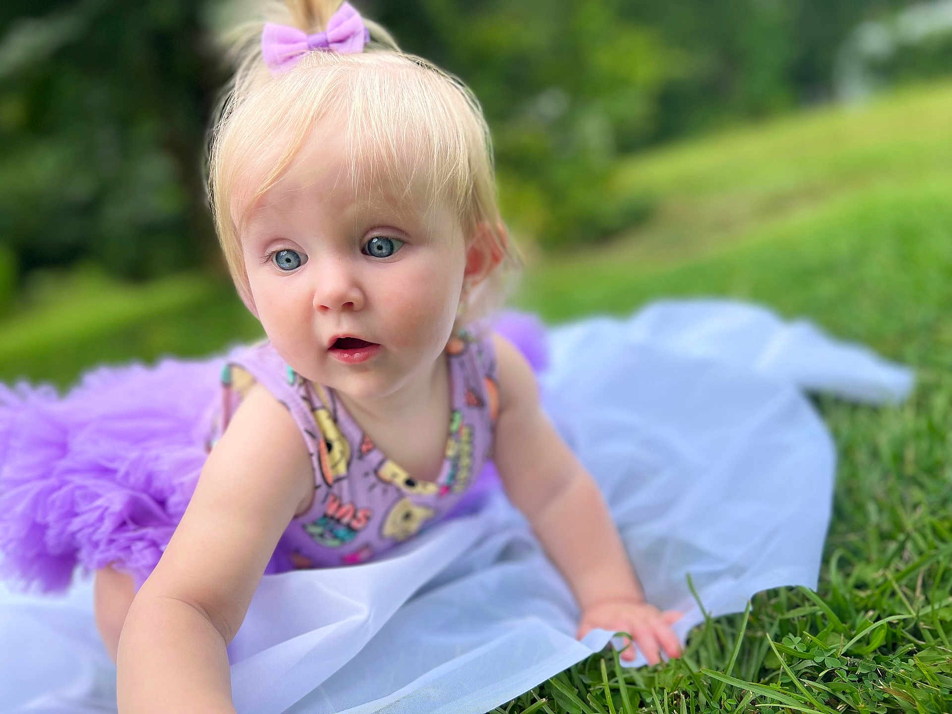 Lila is registered to the contest to win money with this photo: baby, child, purple_bow, tutu, grass, outdoor, blue_eyes, crawling, white_cloth, cute, curious, soft_focus, nature, greenery, summer, playful, portrait, infant, colorful_clothing, daylight