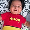 baby, infant, child, smiling, lying_down, portrait, red_clothing, yellow_clothing, onesie, blanket, bed, indoors, phone_screenshot, video_call, face, hair, happy, patterned_background, casual_photo, close_up