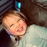 Maëlle participe au concours pour gagner de l'argent avec cette photo : casual_clothing, cheerful, child, close_up, couch, cozy, expression, face, furniture, hair, happy, indoor, light_blue_shirt, person, portrait, skin, smile, text_on_clothing, toddler, young_child