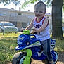 toddler, child, toy_bike, outdoor, smiling, grass, tree, sneakers, shorts, tank_top, sunlight, play, fun, daylight, fence, car, park, happy, young_child, casual_clothing