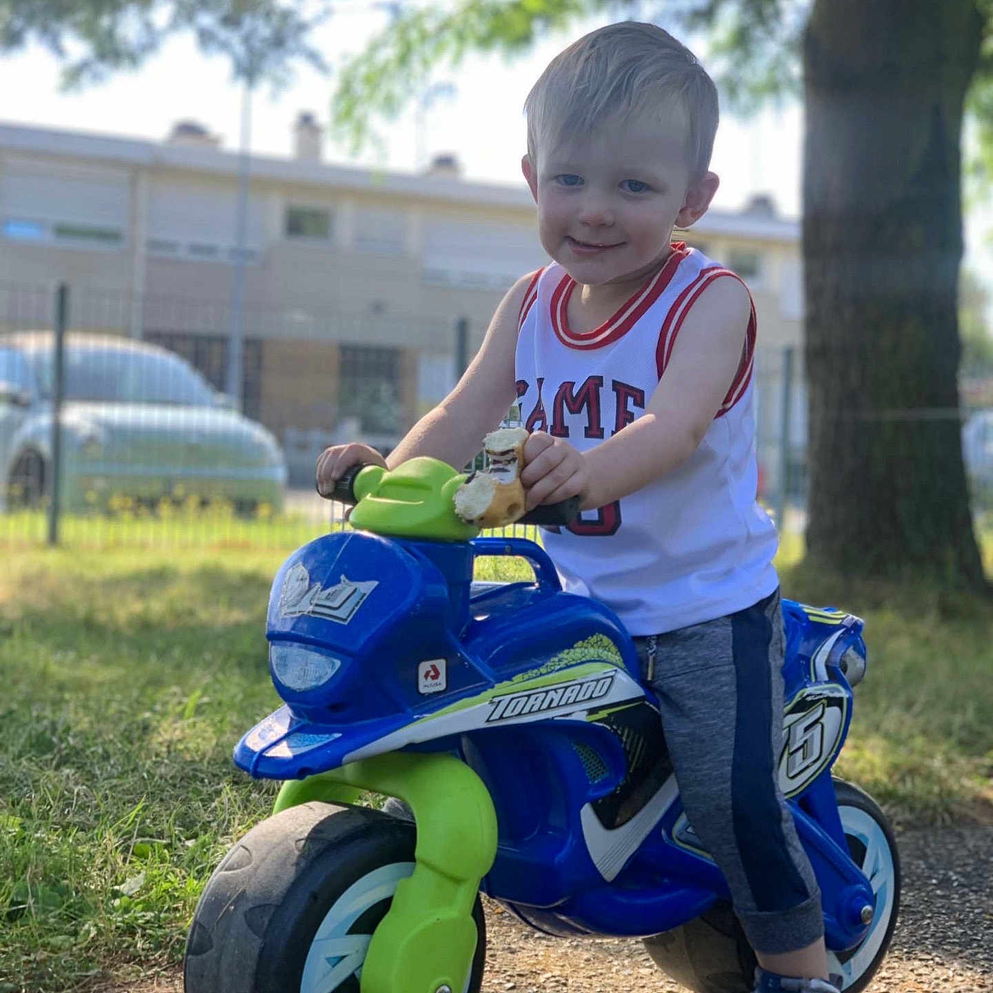 Liam participe au concours pour gagner de l'argent avec cette photo : car, casual_clothing, child, daylight, fence, fun, grass, happy, outdoor, park, play, shorts, smiling, sneakers, sunlight, tank_top, toddler, toy_bike, tree, young_child