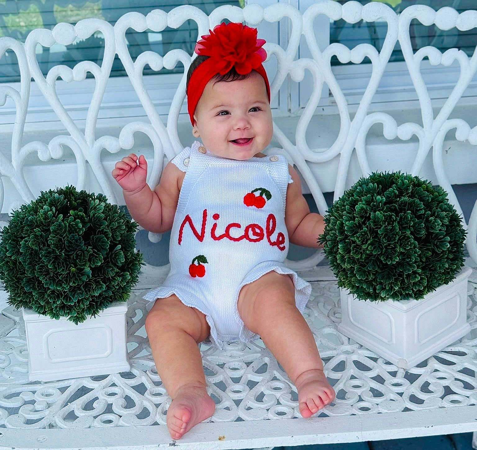 Nicole joined the competition — help win amazing prizes! baby, child, smiling, white_bench, green_plants, potted_plants, red_headband, romper, embroidered_clothing, outdoor, happy, cute, person, infant, sitting, decorative_furniture, garden, portrait, face, summer