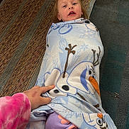 Riki is registered to the contest to win money with this photo: blanket, carpet, casual, child, clothing, comfort, cozy, cute, expression, floor, frozen_olaf, hand, home, indoor, person, playful, purple_pants, toddler, toy, wrapped