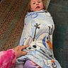 child, blanket, person, carpet, hand, indoor, purple_pants, frozen_olaf, toy, playful, wrapped, cozy, floor, clothing, cute, toddler, expression, casual, home, comfort
