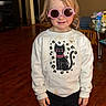child, indoor, pink_sunglasses, white_sweatshirt, black_cat_print, smile, standing, wooden_floor, table, chair, toy_bottle, casual_clothing, long_hair, face, person, home, furniture, light, young, cute