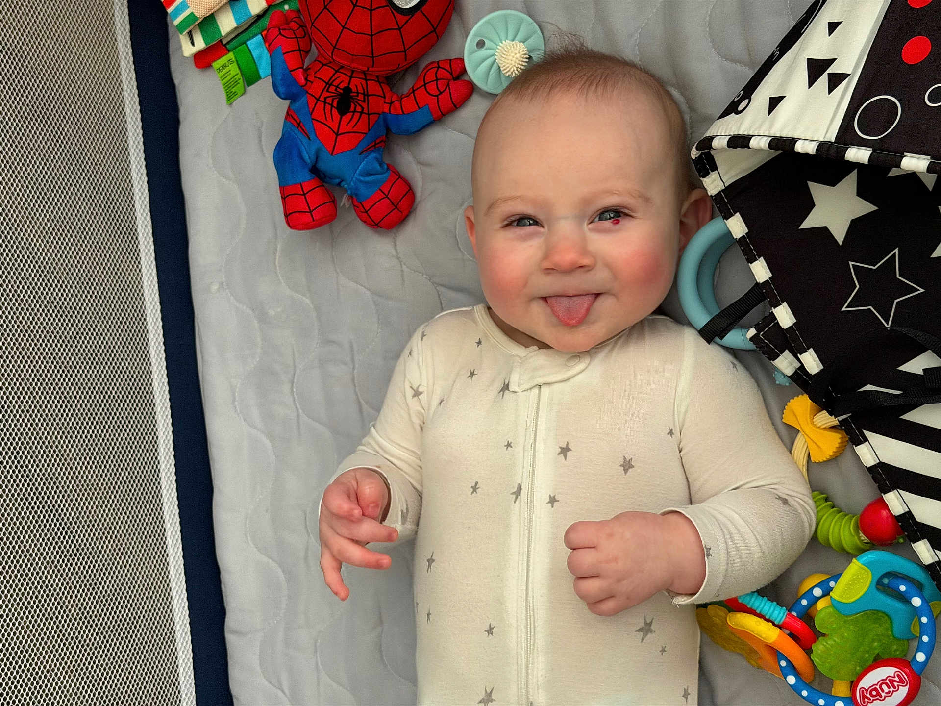 Parker joined the competition — help win amazing prizes! baby, infant, smiling, sticking_tongue, onesie, toys, playpen, spiderman_plush, plush_toy, teether, colorful, play_mat, star_pattern, rosy_cheeks, crib_mesh, lying_down, close_up, portrait, happy, chubby_cheeks